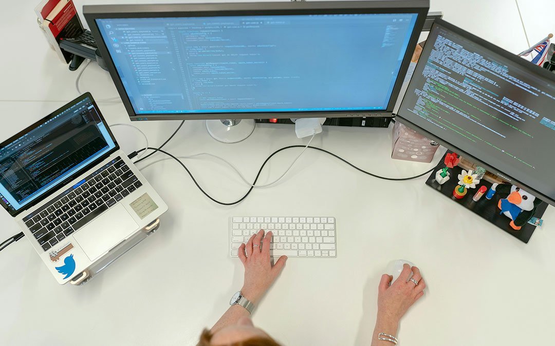 Overhead view of a desk with a laptop and two monitors showing code, with a person's hands at the keyboard and mouse
